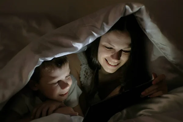 Молодая женщина мама и ее сын смотрят интересный фильм вместе на планшете под одеялом . — стоковое фото