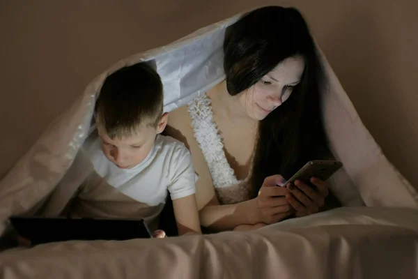 Молодая красивая женщина мама просматривает интернет в своем телефоне в то время как ее сын смотрит мультфильм на планшете . — стоковое фото