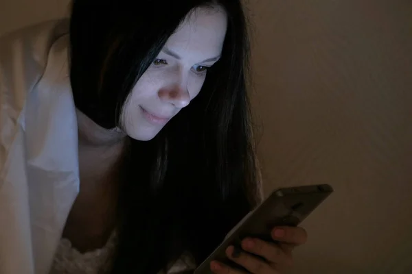 Μελαχρινή νεαρή όμορφη γυναίκα διαβάζει ένα βιβλίο με το κινητό της τηλέφωνο στο κρεβάτι πριν τον ύπνο και χαμόγελα. — Φωτογραφία Αρχείου