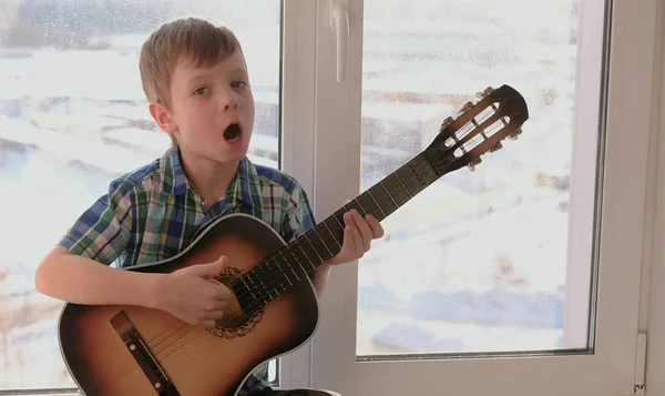 Tocar un instrumento musical. El chico toca la guitarra y canta sentado en el alféizar . — Foto de Stock