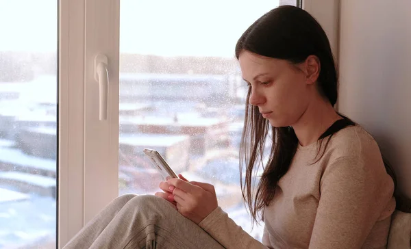 Скучная женщина смотрит что-то в Интернете в своем мобильном телефоне, сидя на подоконнике . — стоковое фото