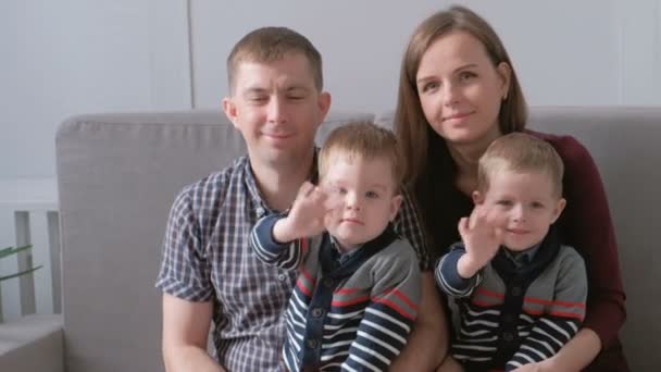 Сімейний відео-портрет мами, тата та двох братів близнюків. Вони сидять на дивані і дивляться на камеру. Хлопці махають руками . — стокове відео