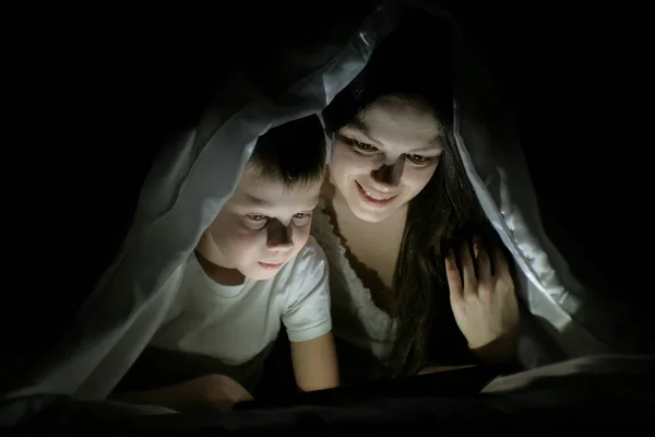 Jovem mãe e seu filho assistindo a um filme juntos em tablet sob o cobertor . — Fotografia de Stock