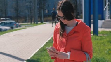 Genç kadın esmer güneş gözlüğü kırmızı paltolu biri için bekler ve onun telefon denetler manifatura.