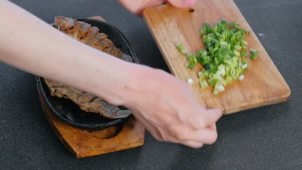 A mulher serve um prato com peixe e cebola verde na cozinha em uma mesa preta. Mãos fechadas . — Vídeo de Stock