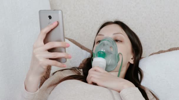 Use nebulizador e inhalador para el tratamiento. Mujer joven inhalando a través de la máscara del inhalador acostada en el sofá y charlando en el teléfono móvil. Vista lateral . — Vídeo de stock