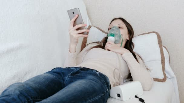 Nebulizatör ve astım tedavisi için kullanın. Genç kadın kanepede yatan ve hareket eden telefon içinde sohbet inhaler maskesi aracılığıyla teneffüs edilmesi. Yan görünüm. — Stok video