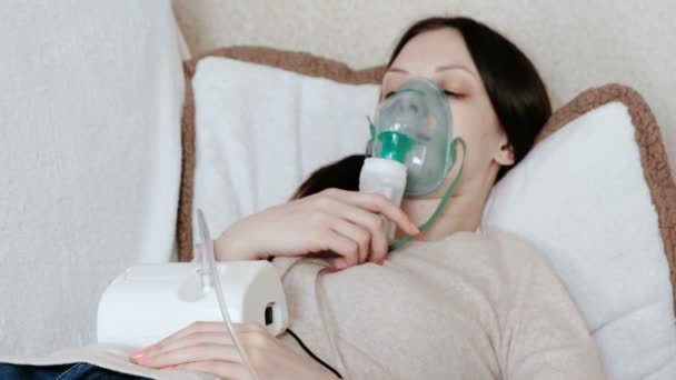 Use nebulizador e inhalador para el tratamiento. Mujer joven inhalando a través de la máscara del inhalador acostada en el sofá. Vista lateral . — Vídeos de Stock