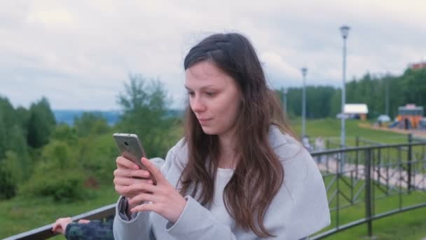 Jonge brunette vrouw chatten, het typen van een bericht op mobiele telefoon wandelen langs de promenade in de stad. — Stockvideo