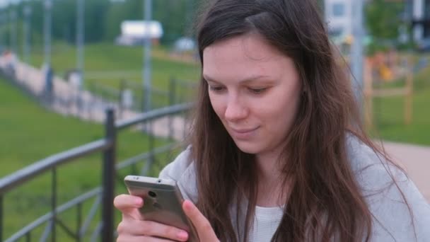 若いブルネットの女性都市の遊歩道に沿って歩いて携帯電話でメッセージを入力して、チャット. — ストック動画
