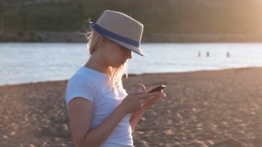 Şapkalı güzel sarışın kız sahilde cep telefonunda günbatımında bir ileti türleri.