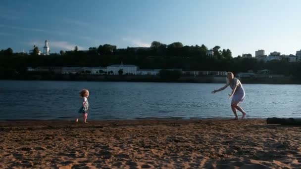 Linda loira mãe e filha correr para o outro na margem do rio ao pôr do sol . — Vídeo de Stock