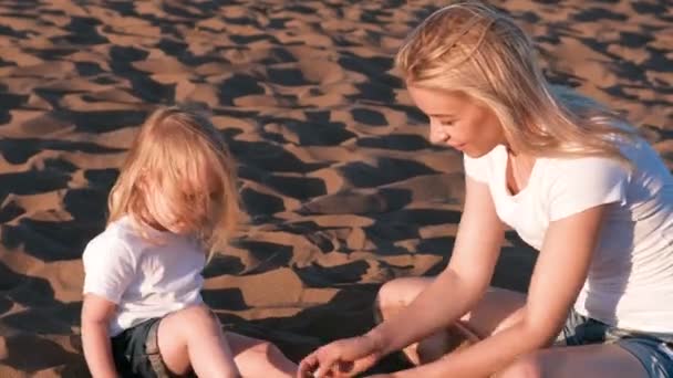 Beautiful blonde mom and daughter play with sand sitting together on the beach. — Stock Video