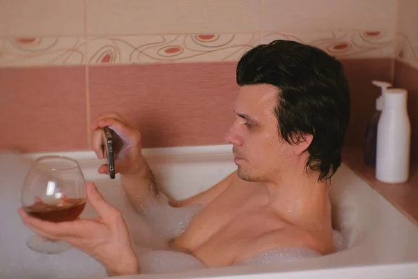 Молодой человек принимает ванну с пеной, смотрит видео по мобильному телефону, пьет виски и улыбается . — стоковое фото