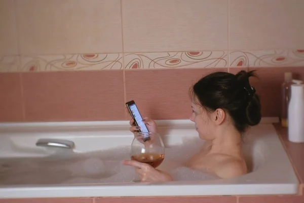 Genç güzel esmer kadın alır banyo, içki viski ve online alışveriş üstünde hareket eden telefon. — Stok fotoğraf