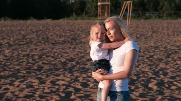 Schöne blonde Mutter und Tochter kuscheln und sprechen am Strand bei Sonnenuntergang. — Stockvideo