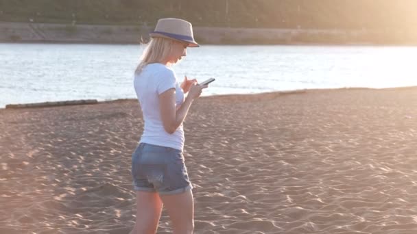 Hermosa chica rubia en un sombrero escribe un mensaje en su teléfono móvil caminando por la playa al atardecer . — Vídeo de stock