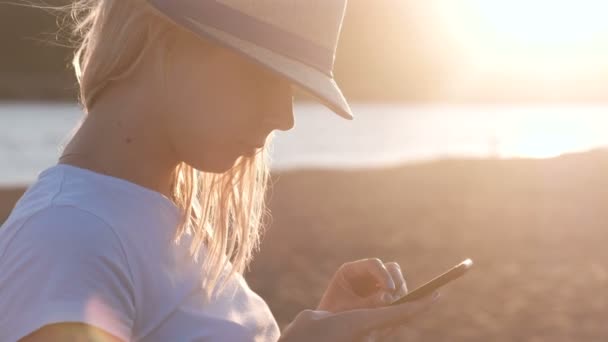 Hermosa chica rubia en un sombrero escribe un mensaje en su teléfono móvil en la playa al atardecer . — Vídeo de stock