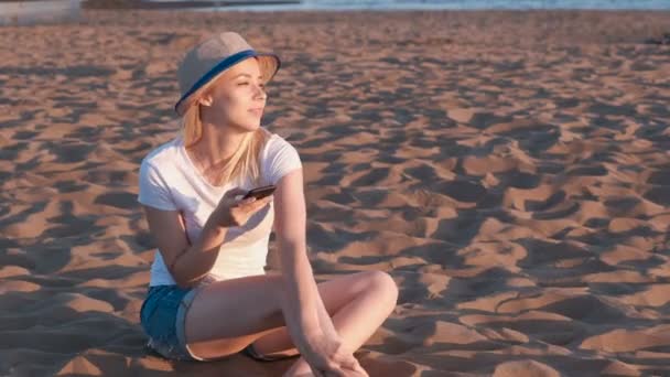 Vacker blond flicka i en hatt som sitter på stranden vid solnedgången med mobiltelefon. — Stockvideo