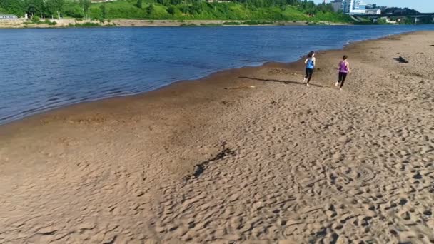 Две женщины бегают по песчаному пляжу реки на закате. Вид на город . — стоковое видео