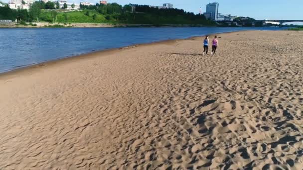 İki kadın nehir kumlu plaj boyunca gün batımında koşu. Güzel şehir manzaralı. — Stok video
