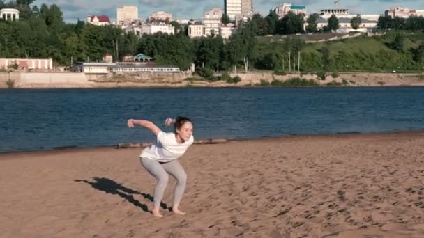 Kvinnan gör övningar sport på stranden av floden i staden. Hoppa från sittande ställning. — Stockvideo