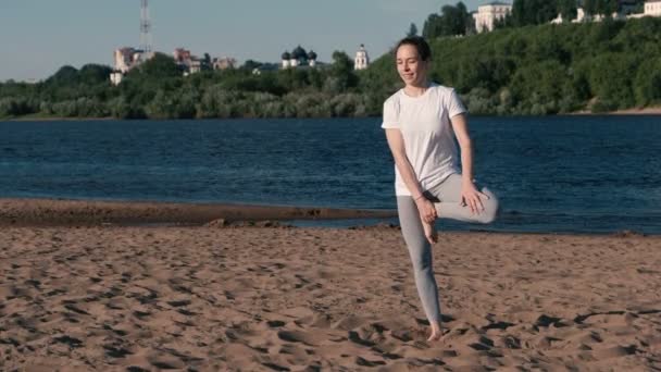 Vrouw die zich uitstrekt van yoga op het strand aan de rivier in de stad. Uitzicht op de mooie stad. Vrikshasana pose. — Stockvideo