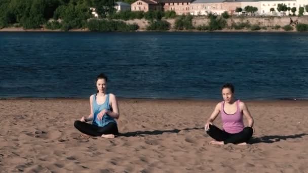 Dos mujeres haciendo yoga en la playa junto al río en la ciudad y hablando. Hermosa vista . — Vídeo de stock