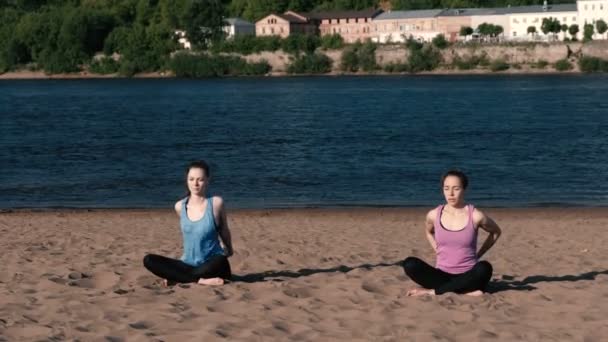 在城市的河边 两个女人在沙滩上伸展瑜伽 美丽的城市景色 冥想姿势 — 图库视频影像