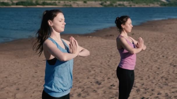 Dos mujeres estirándose yoga de pie en la playa junto al río en la ciudad. Hermosa vista de la ciudad. Postura de Namaste . — Vídeo de stock