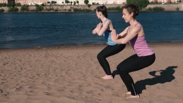 Dos mujeres haciendo yoga en la playa junto al río en la ciudad. Hermosa vista. Utkatasana pose . — Vídeos de Stock