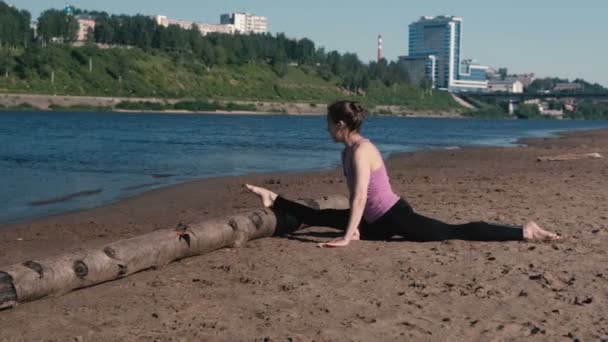 Женщина делает растяжку сидя на бечевке на песчаном пляже на закате. Вид на город . — стоковое видео