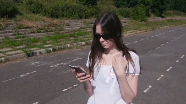 Молода брюнетка дівчина ходить на стадіоні і вводить повідомлення на мобільний телефон і посміхається . — стокове відео