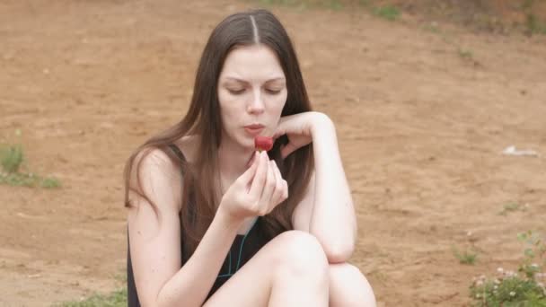 Giovane donna bruna mangia una fragola seduta sulla spiaggia . — Video Stock