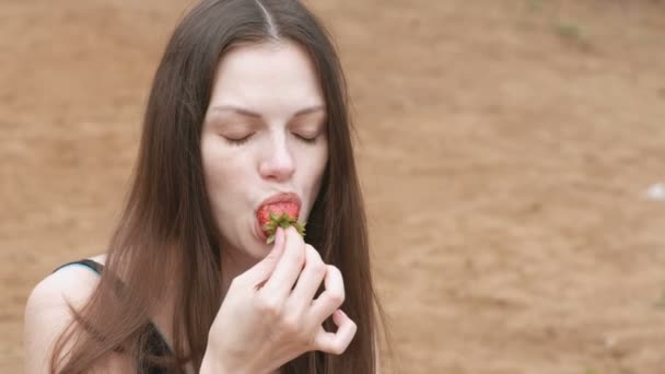 Jeune femme brune mange une fraise assise sur la plage. Gros plan sur le visage . — Video