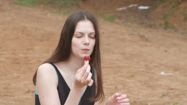 Молода жінка брюнетка їсть полуницю, сидячи на пляжі. Вид збоку . — стокове відео