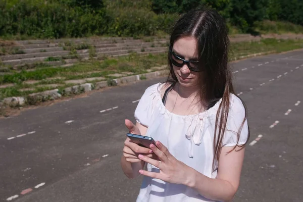 Chica morena joven caminando en el estadio en una zona residencial y mensajería en un teléfono móvil . — Foto de Stock