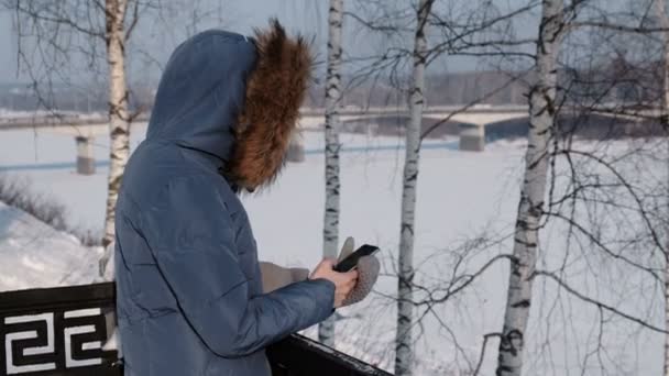 Unrecognizable woman in blue down jacket writes messaging in her cellphone in winter Park. Side view — Stock Video