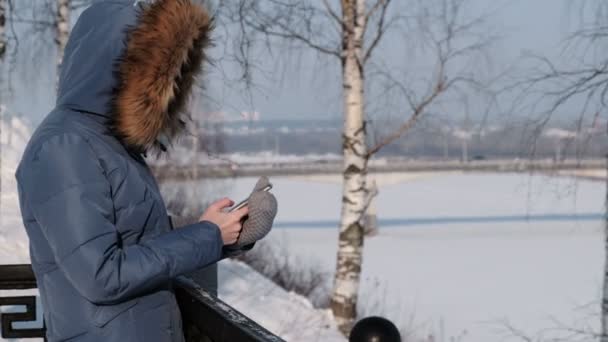 Eine unkenntliche Frau in blauer Daunenjacke schreibt im Winterpark Nachrichten in ihr Handy. Seitenansicht — Stockvideo