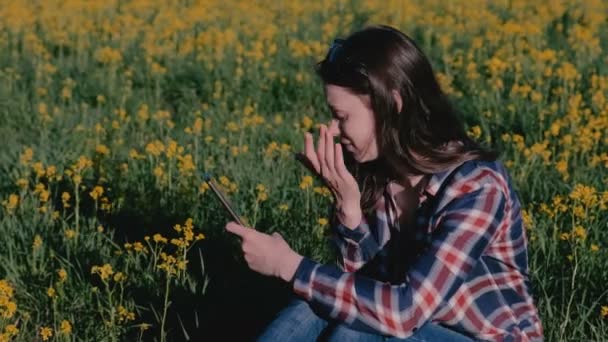 Kobieta, patrząc na telefon komórkowy siedząc w parku na trawie wśród żółte kwiaty. — Wideo stockowe