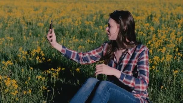 Mulher falar vídeo chat no telefone sentado na grama entre as flores amarelas . — Vídeo de Stock