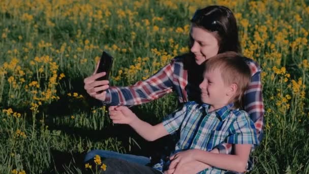 Maman et son fils font selfie au téléphone assis sur l'herbe parmi les fleurs jaunes . — Video