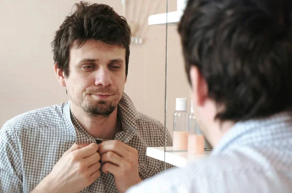 El hombre cansado y somnoliento que acaba de despertarse mira su reflejo en el espejo y ve su apariencia desaliñada, abotonándose con una camisa a cuadros. . —  Fotos de Stock