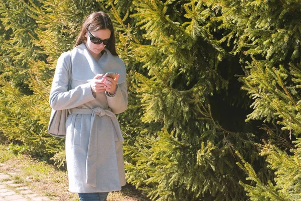 Junge brünette Frau mit Sonnenbrille und Mantel spaziert durch Stadtpark und schaut aufs Handy. — Stockfoto