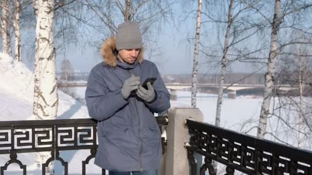 Homem de casaco azul para baixo com capuz de pele pegue seu celular e tira a luva em um parque de inverno . — Vídeo de Stock