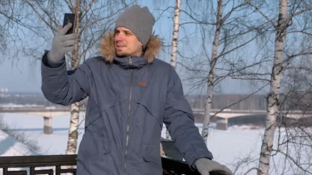 Άνθρωπος με μπλε χρώμα κάτω σακάκι με κουκούλα γούνα που χρησιμοποιούν κινητό για video κουβέντα και χαμογελά στο χειμερινό πάρκο. — Αρχείο Βίντεο