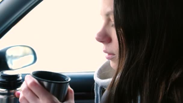Ung brunett kvinna dricker te från en termos som sitter i en bil på vintern. — Stockvideo