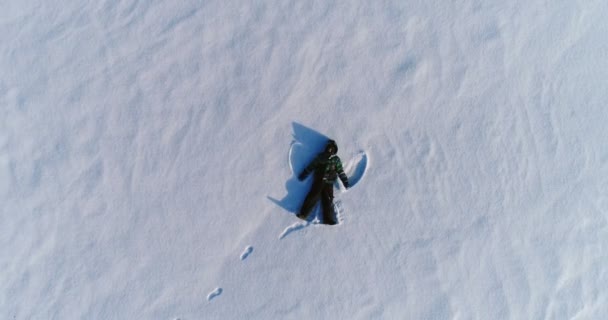 Niño de 7 años en chaqueta azul hace ángel de la nieve, imágenes aéreas. La cámara está subiendo lentamente — Vídeo de stock