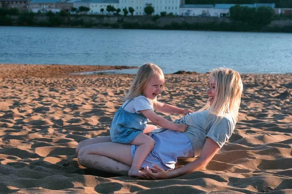 Piękny szczęśliwy Blondynka Mama i Córka, przytulanie i mówiąc, siedząc na plaży o zachodzie słońca. — Zdjęcie stockowe