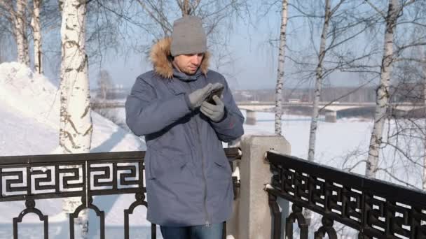 Homme veste bleue avec capuche en fourrure essuie l'écran du téléphone avec sa main dans des gants en hiver Parc . — Video